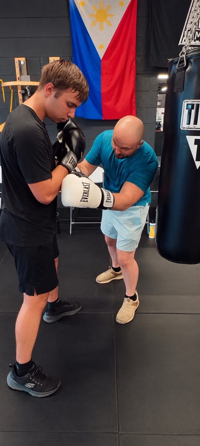 A coach demonstrates positioning to a young boxer wearing gloves beside a heavy bag
