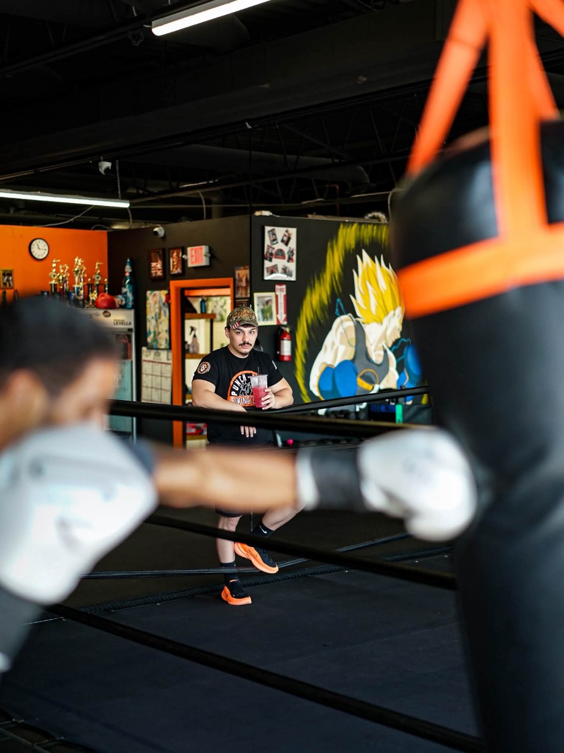 A coach watches from the ring apron while a boxer trains on the heavy bag