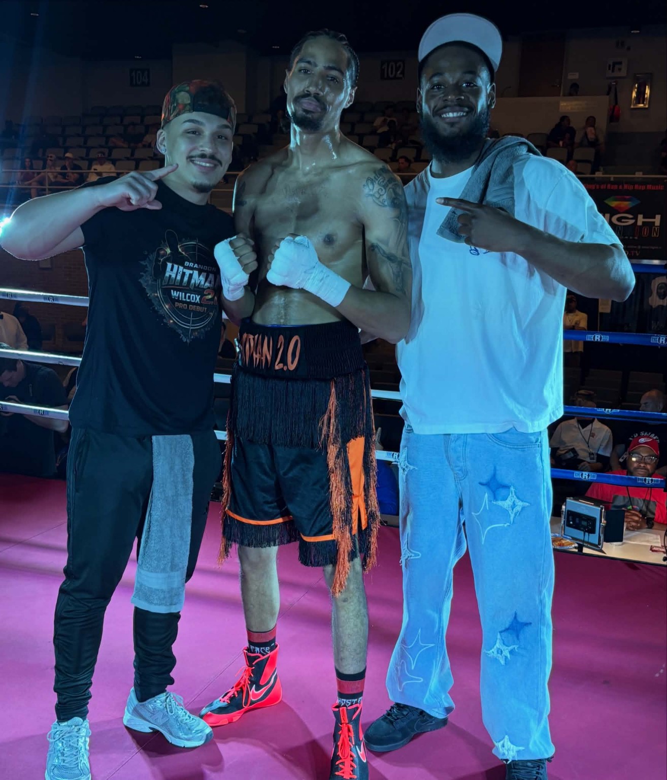 A shirtless boxer in black shorts posing between two teammates in a boxing ring