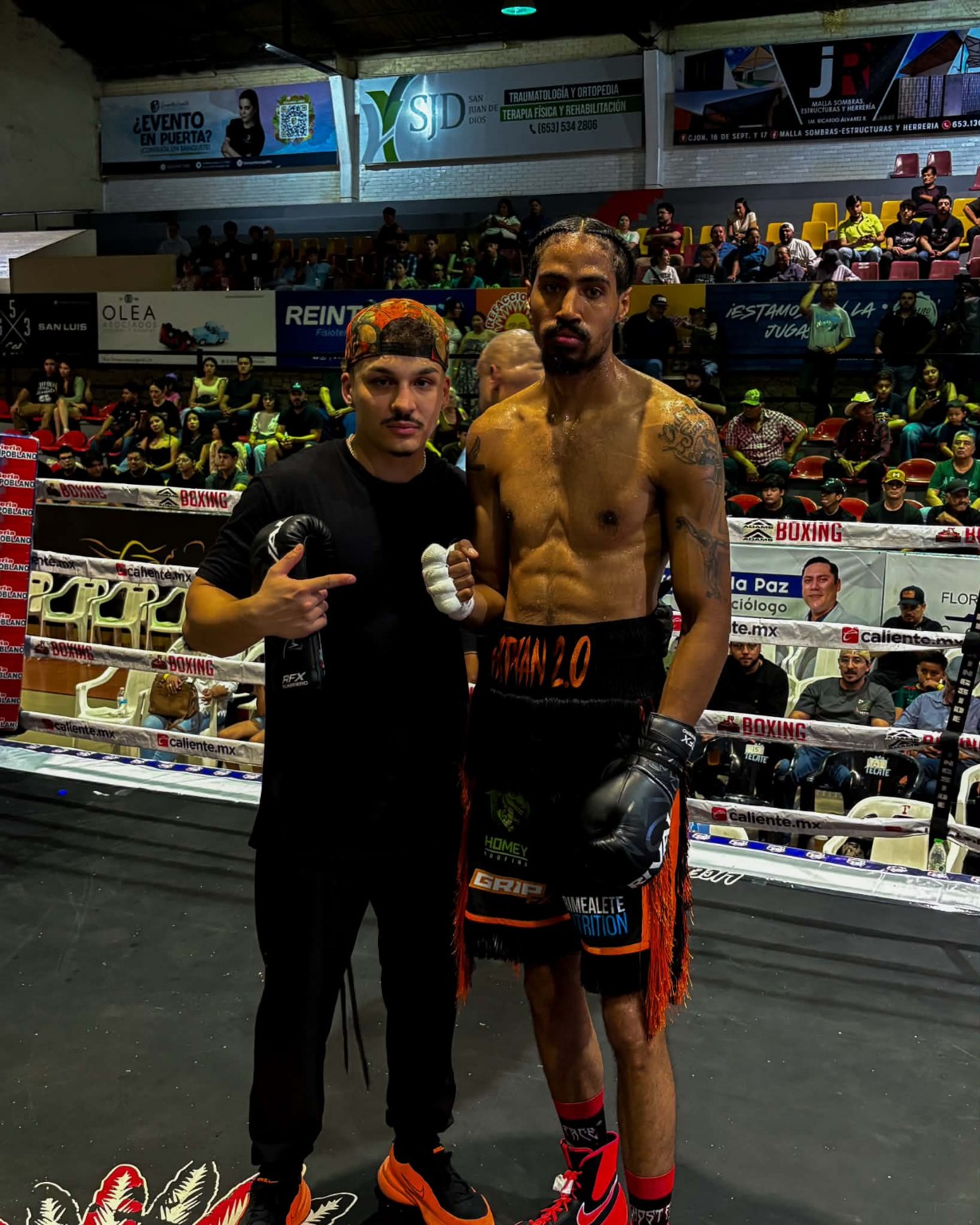 A shirtless boxer in black and orange shorts posing with a coach inside a boxing ring