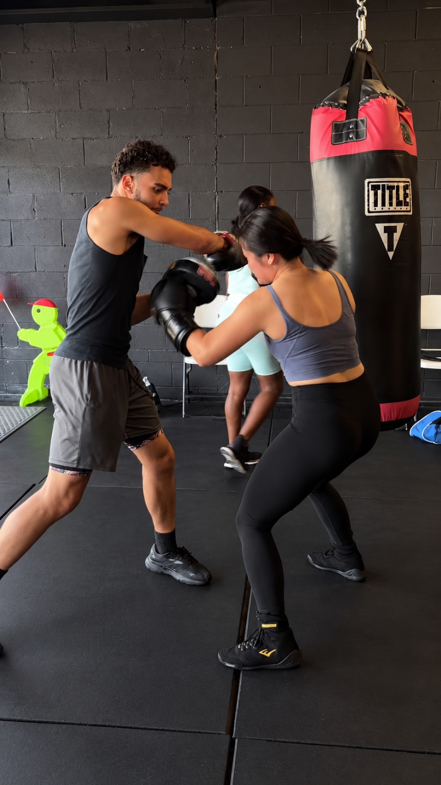 Coach holding focus mitts while a woman boxer practices punches beside a heavy bag