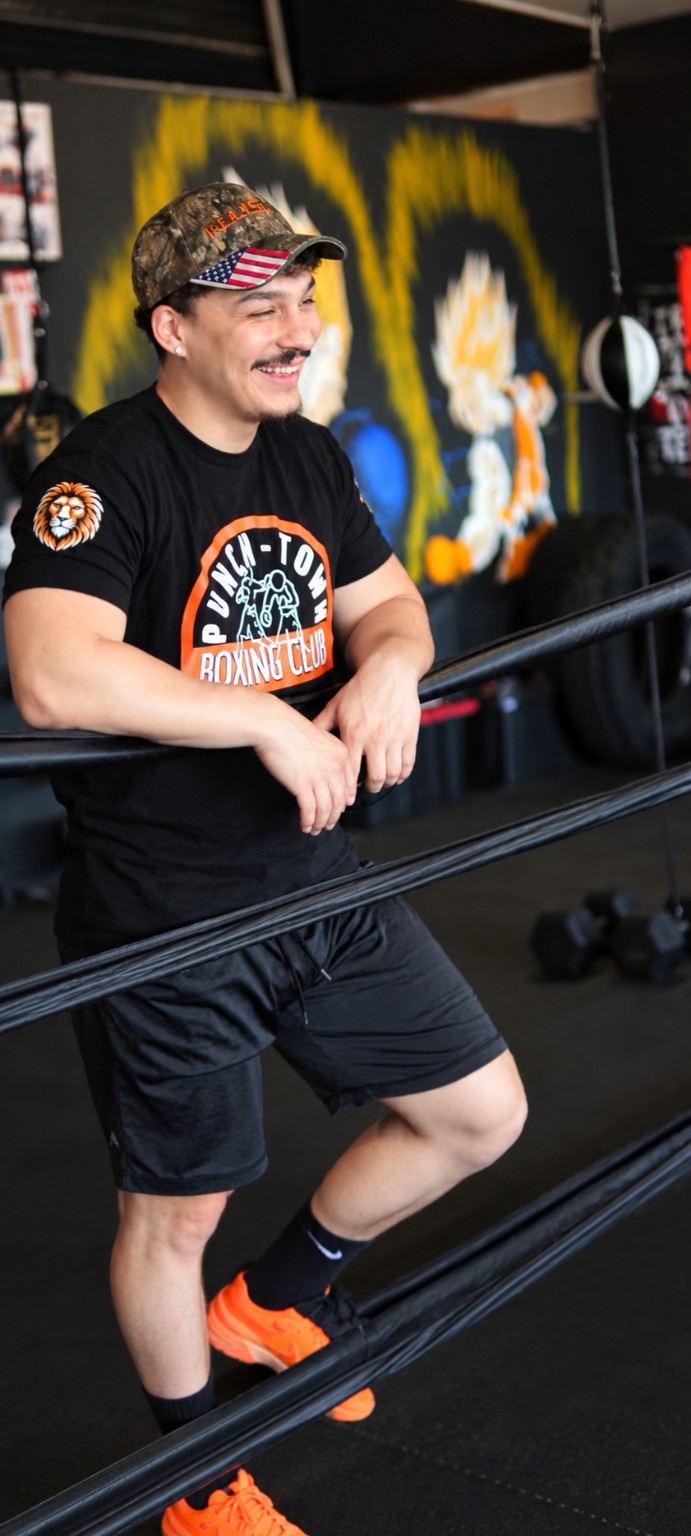 A smiling PunchTown coach in a club t-shirt leans on the ring ropes during youth boxing training